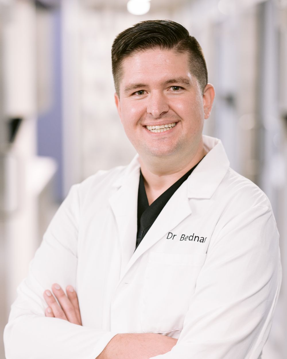 Dr. Josh Bednar smiling with his arms crossed, wearing a white coat over dark scrubs